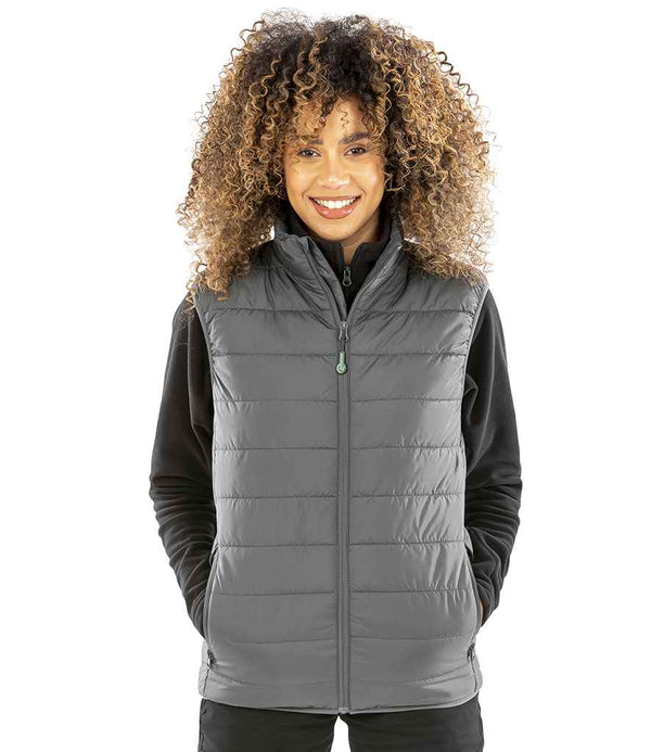 A person with curly hair stands against a white background, smiling at the camera in a gray Result Recycled Promo Padded Bodywarmer over a black long-sleeve top, hands tucked in the vest pockets.