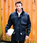 A man wearing the Result Work-Guard Sabre Long Coat by Result Work-Guard stands before an orange shipping container, holding a white hard hat in his left hand and looking at the camera.