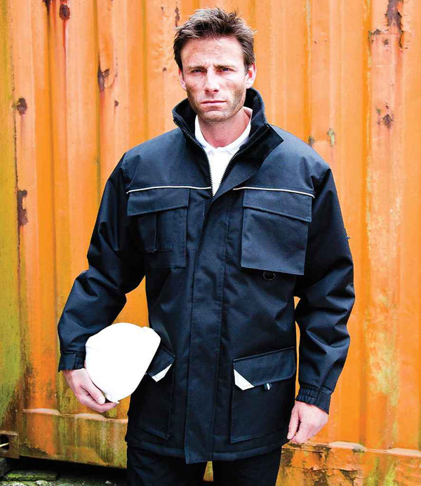 A man wearing the Result Work-Guard Sabre Long Coat by Result Work-Guard stands before an orange shipping container, holding a white hard hat in his left hand and looking at the camera.