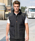 A man stands outdoors on a paved area, wearing the Result Work-Guard Vostex Bodywarmer over a white shirt, with trucks and industrial buildings in the background.