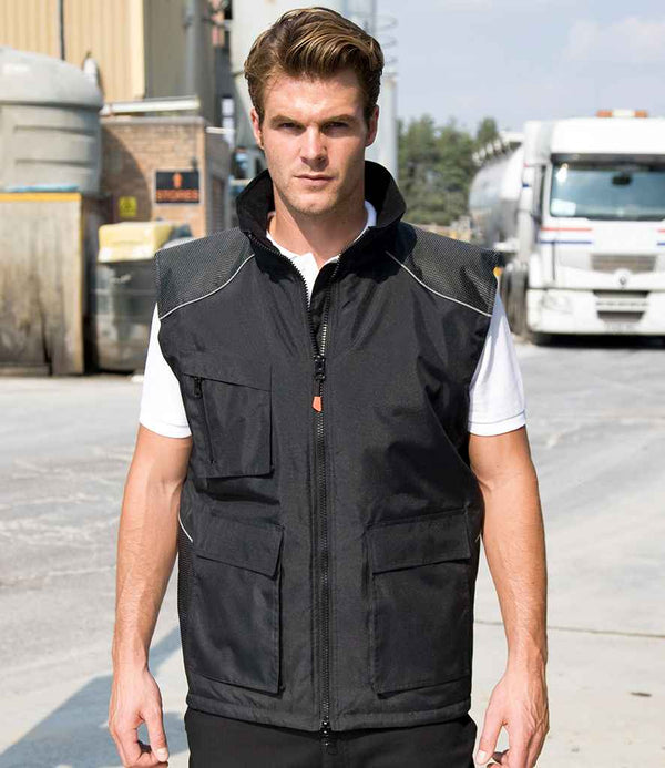 A man stands outdoors on a paved area, wearing the Result Work-Guard Vostex Bodywarmer over a white shirt, with trucks and industrial buildings in the background.