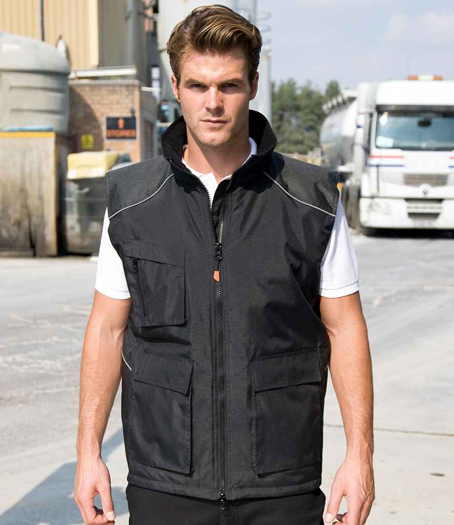 A man stands outdoors on a paved area, wearing the Result Work-Guard Vostex Bodywarmer over a white shirt, with trucks and industrial buildings in the background.