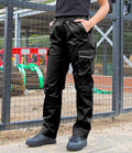 A person wearing Result Work-Guard Ladies Action Trousers and black shoes stands on dirt, hands in pockets, with a construction site featuring fencing and equipment in the background.