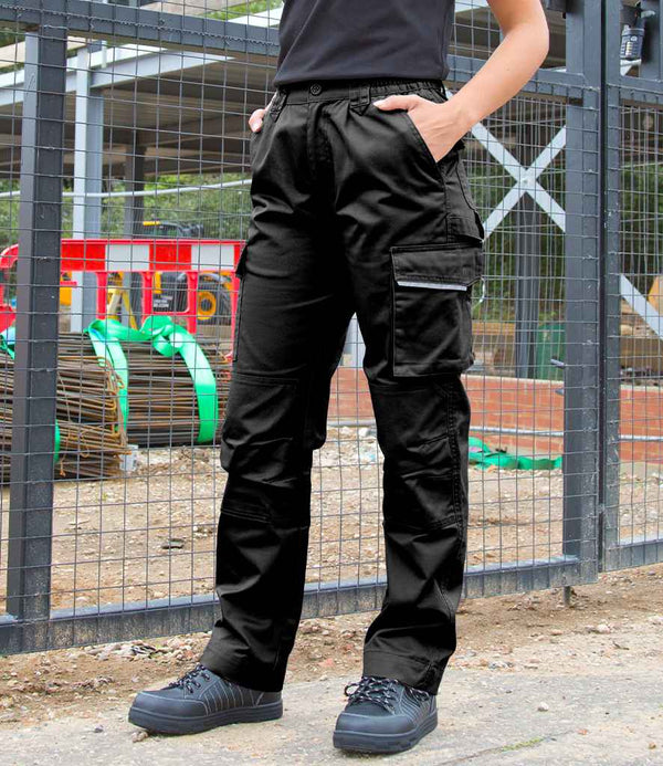 A person wearing Result Work-Guard Ladies Action Trousers and black shoes stands on dirt, hands in pockets, with a construction site featuring fencing and equipment in the background.