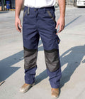 A person in Result Work-Guard Technical Trousers (blue, with black reinforced knee patches and knee pad pockets) by Result Work-Guard, a white T-shirt, and brown work boots stands on an outdoor concrete surface. Their face is not visible.