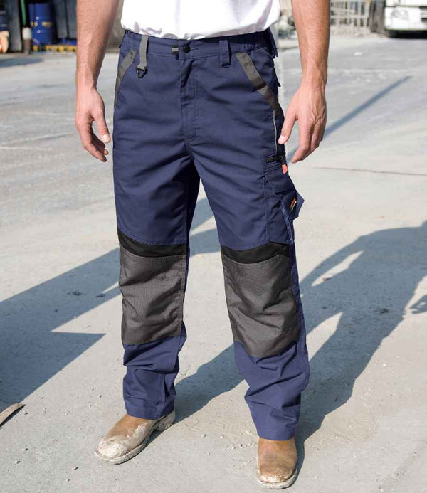 A person in Result Work-Guard Technical Trousers (blue, with black reinforced knee patches and knee pad pockets) by Result Work-Guard, a white T-shirt, and brown work boots stands on an outdoor concrete surface. Their face is not visible.
