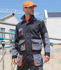 A man in a black and gray Result Work-Guard Lite Jacket by Result Work-Guard, featuring multiple pockets and orange accents, stands at a construction site wearing an orange baseball cap.
