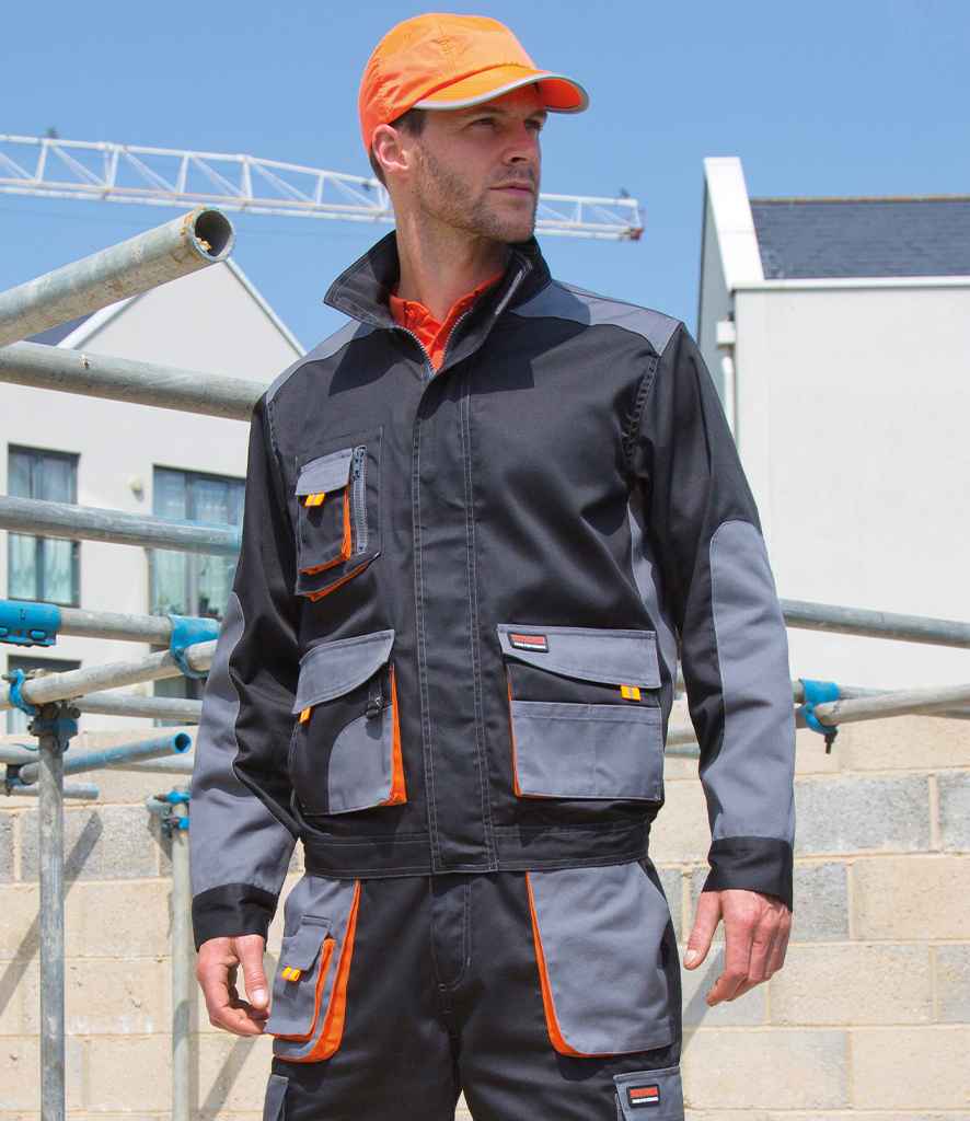 A man in a black and gray Result Work-Guard Lite Jacket by Result Work-Guard, featuring multiple pockets and orange accents, stands at a construction site wearing an orange baseball cap.
