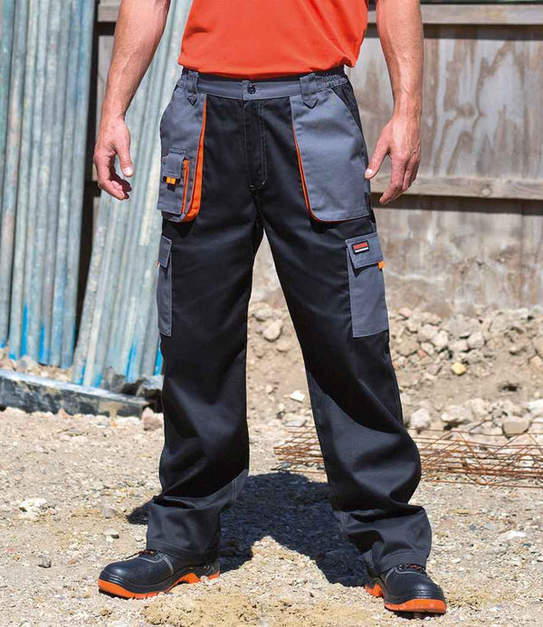 Outdoors on gravel, a person wears Result Work-Guard Lite Trousers by Result Work-Guard—black and gray with orange accents, windproof fabric, and cargo pockets—paired with an orange shirt and black work shoes near a wooden fence and construction materials.