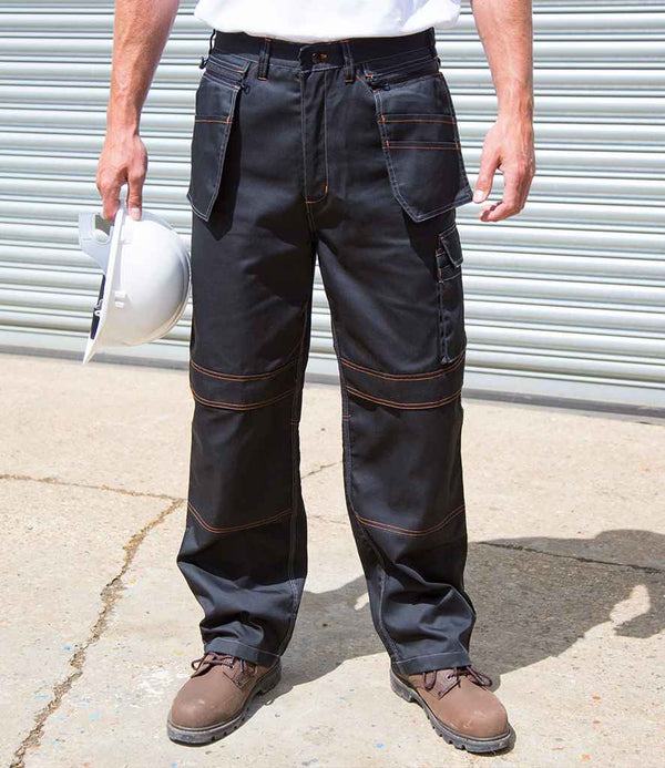 A person in Result Work-Guard Lite X Over Holster Trousers—black with orange stitching and knee pad pockets—brown work boots, and a white shirt stands on concrete holding a white hard hat. A metal roll-up door is behind them.