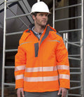 A man wearing a white hard hat and a Result Safe-Guard Dynamic Soft Shell Jacket with reflective stripes stands in front of metal scaffolding at a construction site.