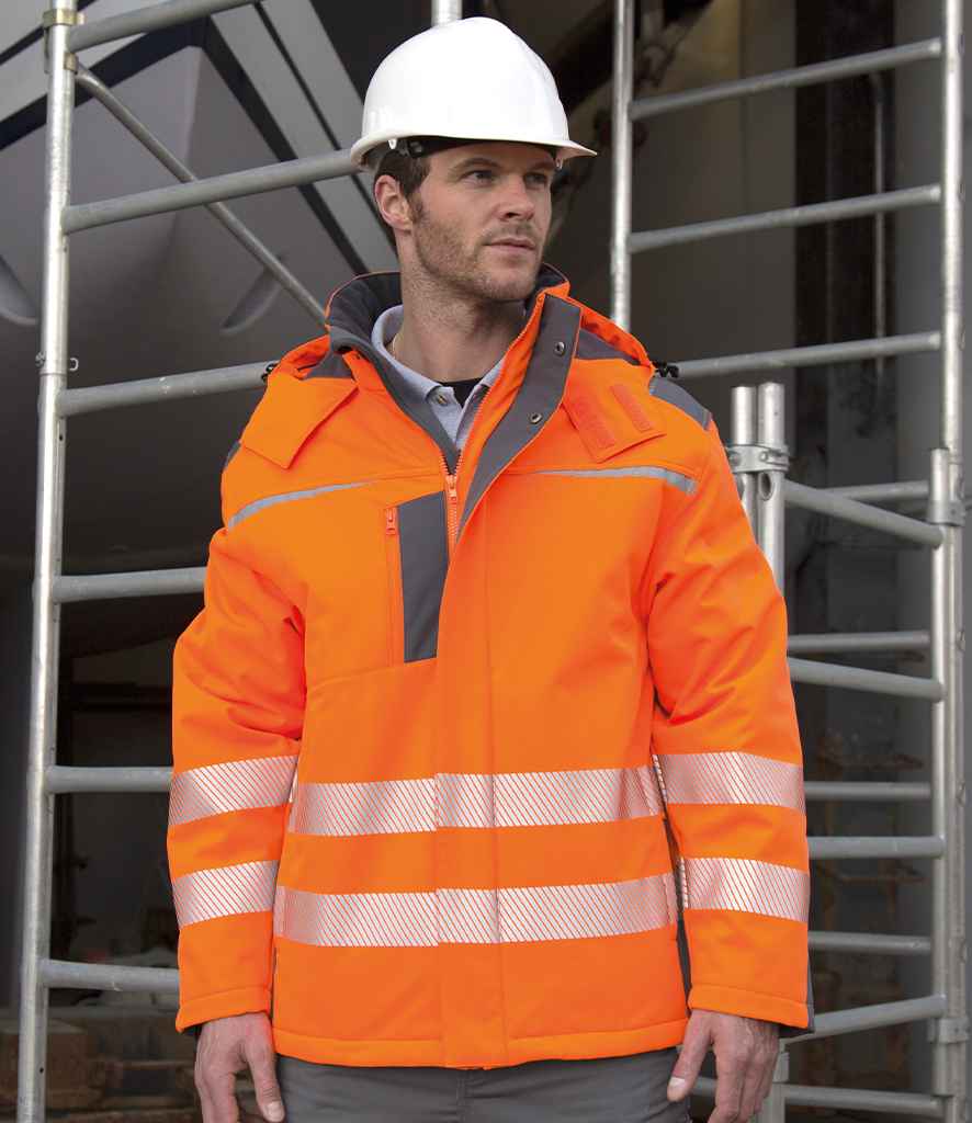 A man wearing a white hard hat and a Result Safe-Guard Dynamic Soft Shell Jacket with reflective stripes stands in front of metal scaffolding at a construction site.