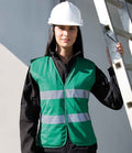 A person wearing a white hard hat and a Result Core Ladies Hi-Vis Vest in green with reflective bands stands next to a metal ladder, holding it with one hand. The background is plain and light-colored.