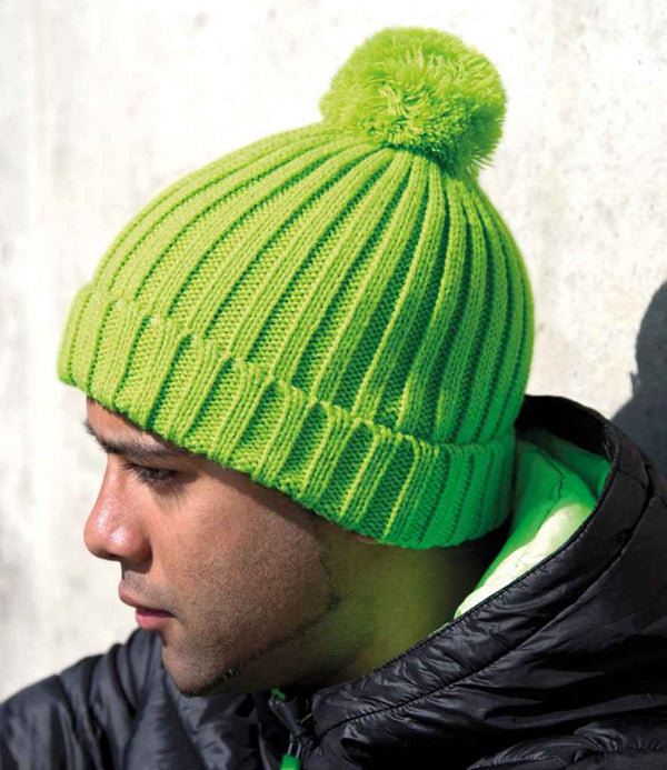 A person in profile, wearing the Result Winter Essentials HDi Quest Knitted Hat—a bright green, windproof beanie with a ribbed knit and pom-pom—paired with a black jacket against a light background.