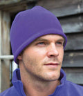 A man wears a Result Winter Essentials Result Reversible Fleece Skull Hat in purple with a matching jacket, gazing slightly to the side. Behind him are weathered wooden boards and a window.