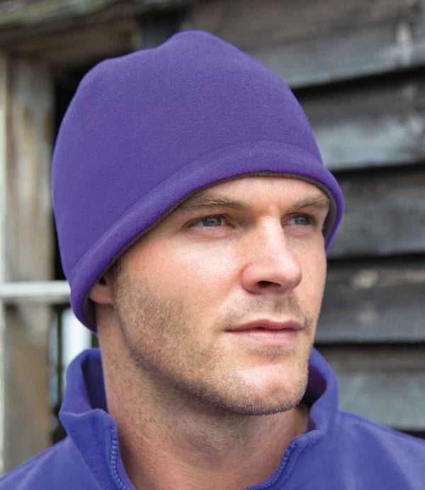 A man wears a Result Winter Essentials Result Reversible Fleece Skull Hat in purple with a matching jacket, gazing slightly to the side. Behind him are weathered wooden boards and a window.