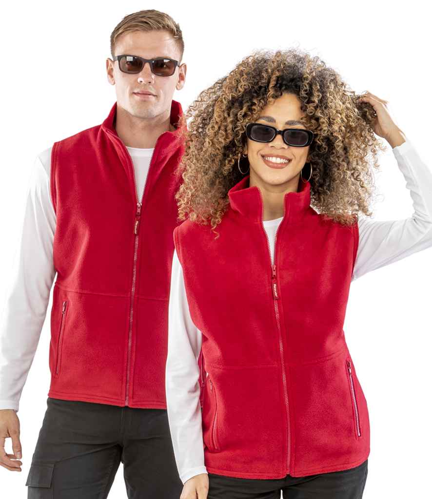 A man and a woman, both smiling and wearing sunglasses, white long-sleeve shirts, and red Result Polartherm™ Fleece Bodywarmers by Result. The woman with curly hair touches her head as the man stands slightly behind her.