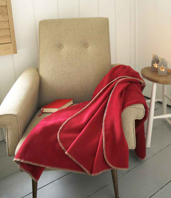 A beige, pill-resistant armchair with a Result Winter Essentials Result Polartherm™ Blanket in red draped over one arm and a closed red book on the seat. A round side table holds two house-shaped candle holders with lit candles.