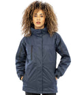 A woman with curly hair stands facing forward against a plain background, hands in the pockets of a dark blue Result Ladies Journey 3-in-1 Jacket with Soft Shell Inner by Result, featuring a high collar and zippered pockets.