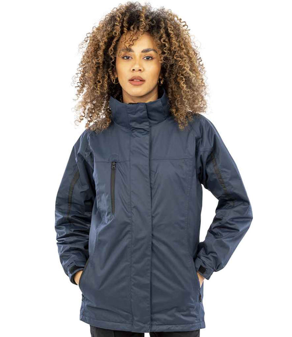 A woman with curly hair stands facing forward against a plain background, hands in the pockets of a dark blue Result Ladies Journey 3-in-1 Jacket with Soft Shell Inner by Result, featuring a high collar and zippered pockets.