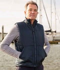 A man stands outdoors by water and boats, facing the camera with a neutral expression. He wears a gray long-sleeve shirt and the Result Fleece Lined Bodywarmer by Result, keeping his hands in the vest pockets.