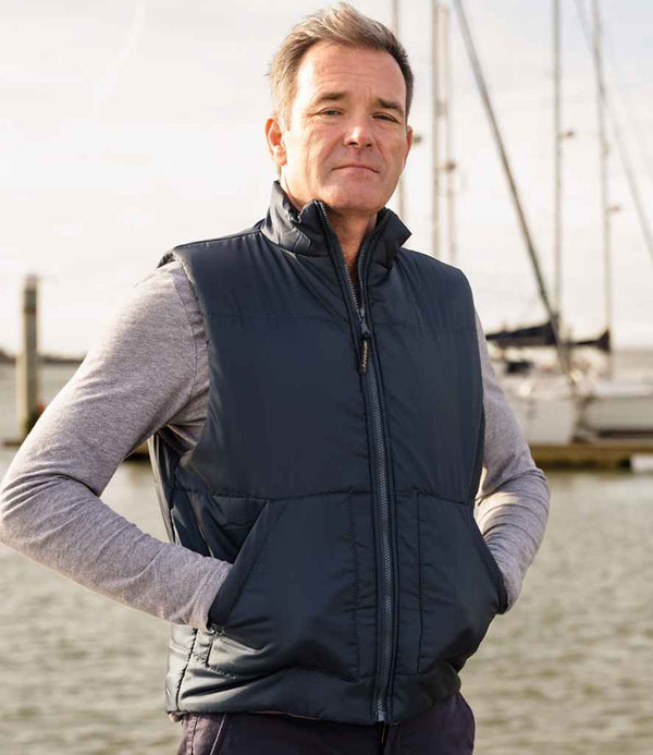 A man stands outdoors by water and boats, facing the camera with a neutral expression. He wears a gray long-sleeve shirt and the Result Fleece Lined Bodywarmer by Result, keeping his hands in the vest pockets.