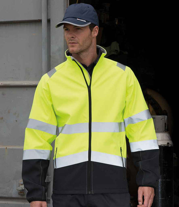 A man wears a Result Safe-Guard Printable Safety Soft Shell Jacket by Result Safe-Guard, featuring high-visibility yellow and black panels and EN ISO 20471 certification, along with a black cap and pants, standing before an open industrial doorway.