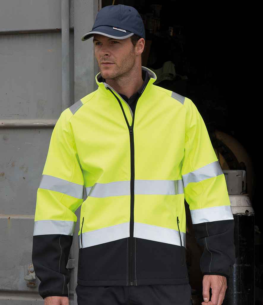 A man wears a Result Safe-Guard Printable Safety Soft Shell Jacket by Result Safe-Guard, featuring high-visibility yellow and black panels and EN ISO 20471 certification, along with a black cap and pants, standing before an open industrial doorway.