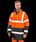 A man stands facing forward in a Result Core Motorway Two Tone Safety Jacket by Result Core—an orange and black waterproof hi vis jacket with white reflective stripes, worn over orange pants. The jacket meets EN ISO 20471 standards. The background is solid black.