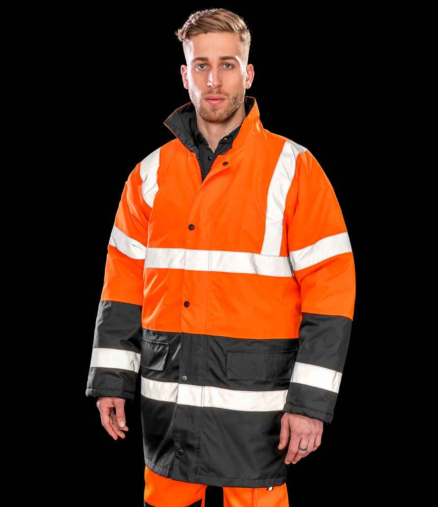 A man stands facing forward in a Result Core Motorway Two Tone Safety Jacket by Result Core—an orange and black waterproof hi vis jacket with white reflective stripes, worn over orange pants. The jacket meets EN ISO 20471 standards. The background is solid black.