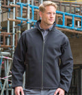 A man wearing the Result Work-Guard Treble Stitch Soft Shell Jacket by Result Work-Guard over a grey collared shirt stands outdoors in front of a building under construction with visible scaffolding.