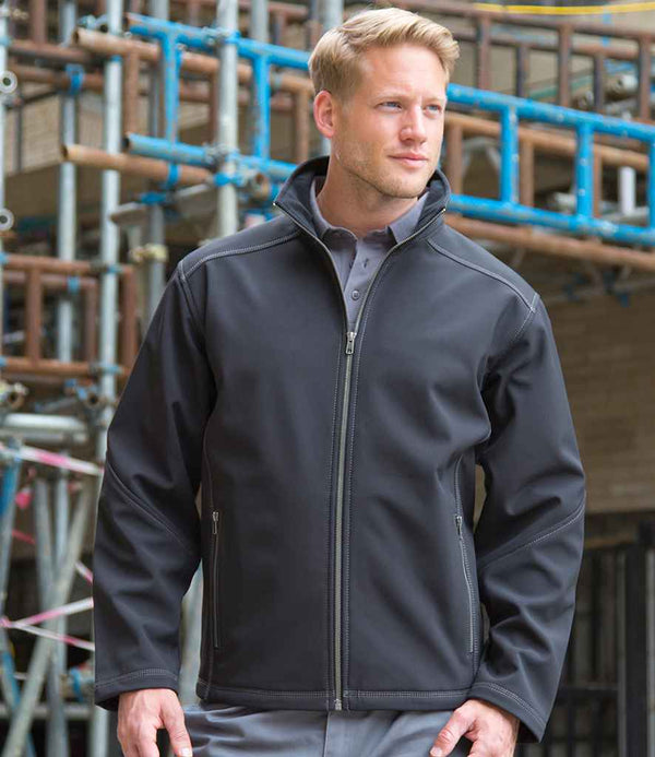 A man wearing the Result Work-Guard Treble Stitch Soft Shell Jacket by Result Work-Guard over a grey collared shirt stands outdoors in front of a building under construction with visible scaffolding.