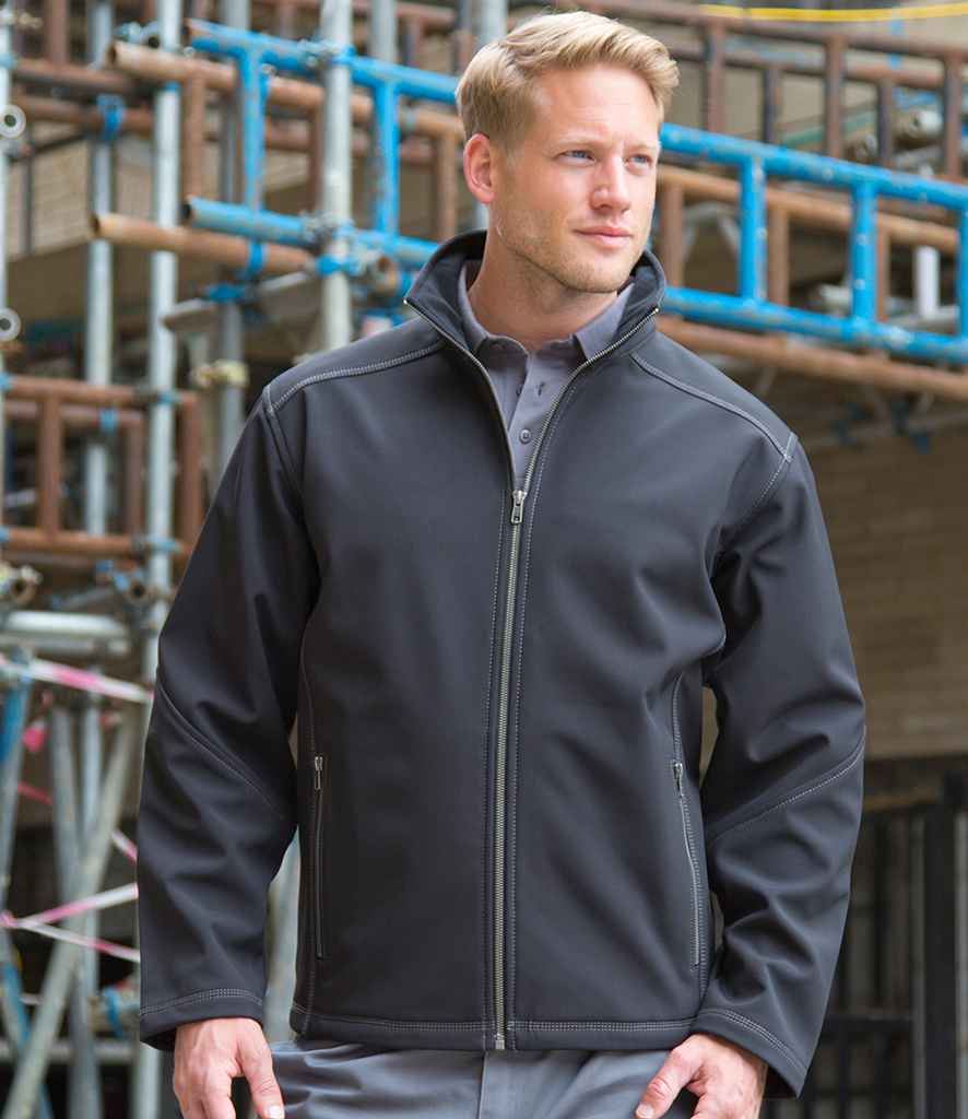 A man wearing the Result Work-Guard Treble Stitch Soft Shell Jacket by Result Work-Guard over a grey collared shirt stands outdoors in front of a building under construction with visible scaffolding.