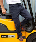 Someone wearing Result Work-Guard Super Stretch Slim Chino Trousers and orange sneakers stands on the step of a yellow forklift marked 15L - 7M, with only their lower half visible against a corrugated metal wall.
