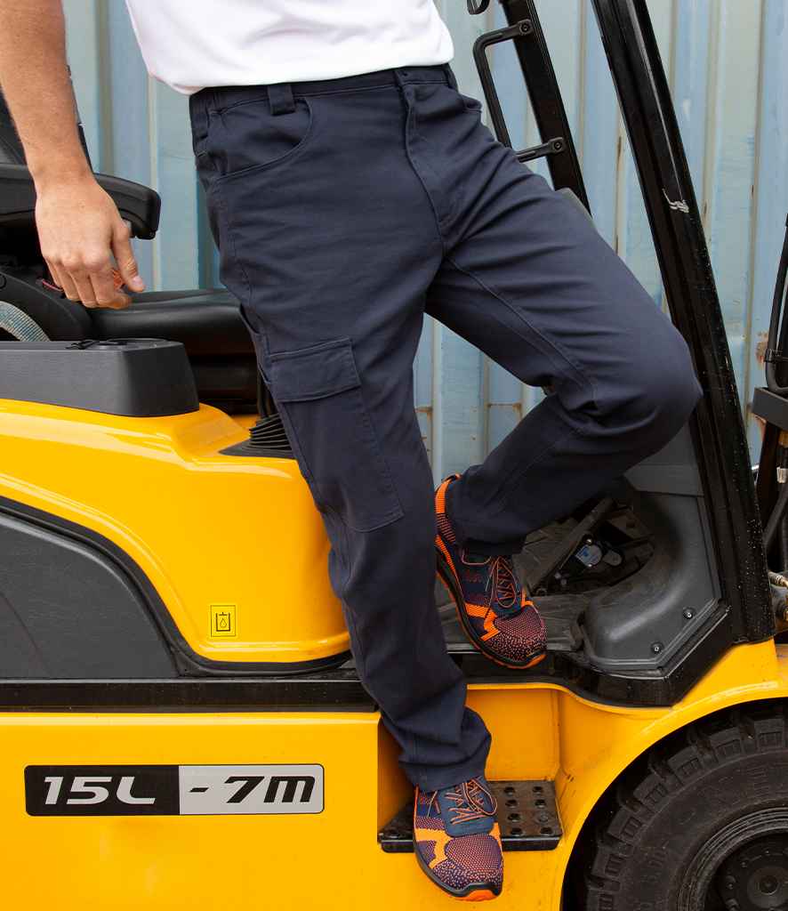 Someone wearing Result Work-Guard Super Stretch Slim Chino Trousers and orange sneakers stands on the step of a yellow forklift marked 15L - 7M, with only their lower half visible against a corrugated metal wall.