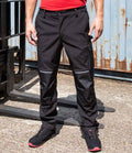 A person wearing Result Work-Guard Slim Fit Soft Shell Trousers, a red shirt, and black work shoes stands on concrete near industrial equipment and a rusted metal wall.