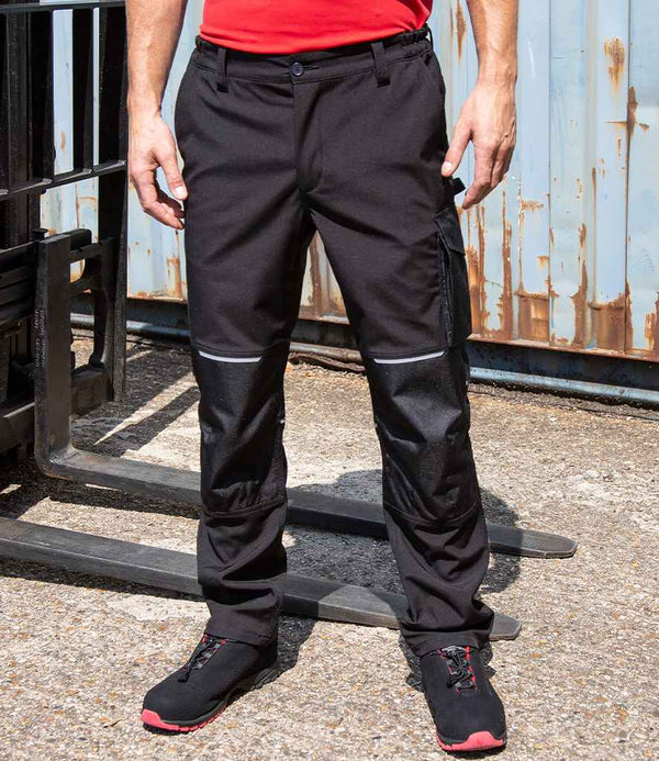 A person wearing Result Work-Guard Slim Fit Soft Shell Trousers, a red shirt, and black work shoes stands on concrete near industrial equipment and a rusted metal wall.
