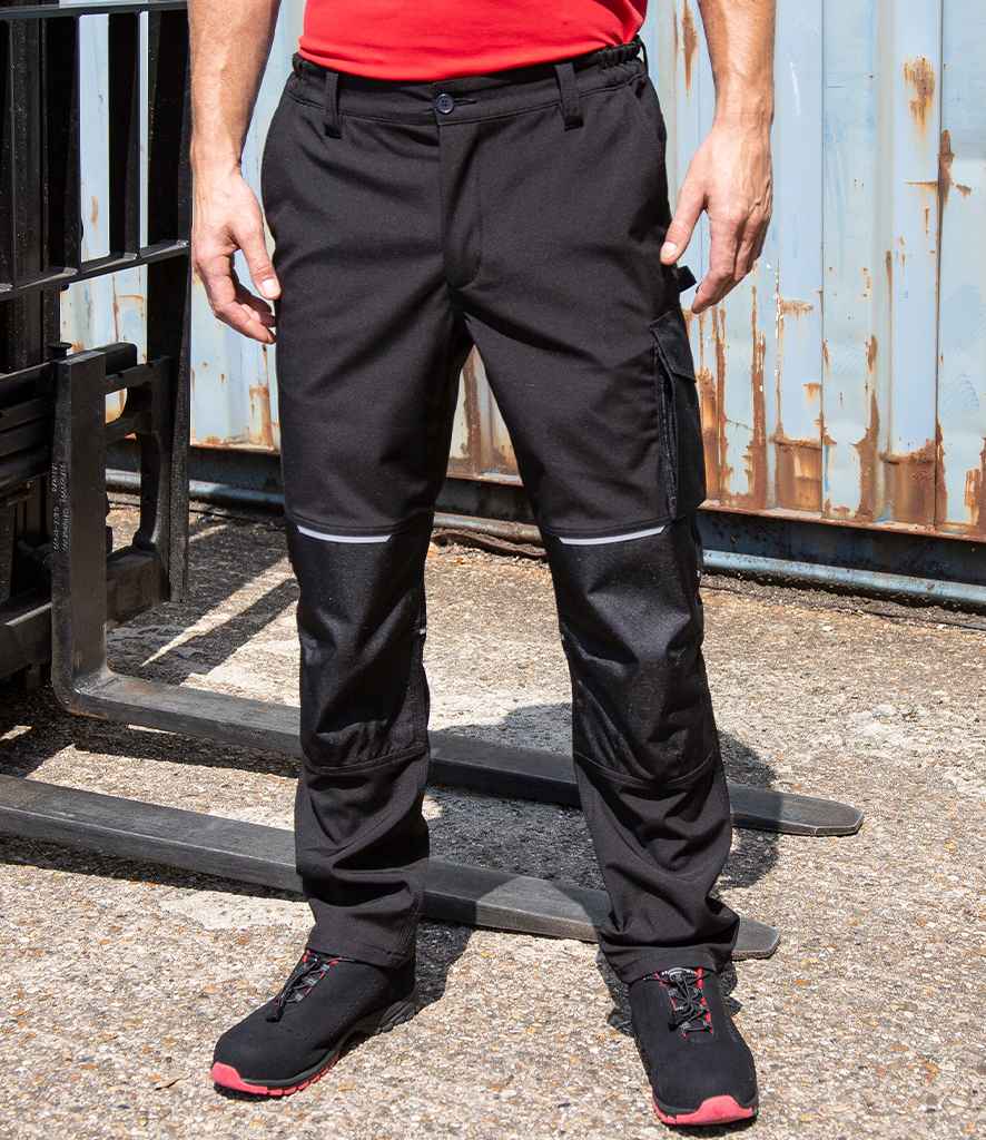 A person wearing Result Work-Guard Slim Fit Soft Shell Trousers, a red shirt, and black work shoes stands on concrete near industrial equipment and a rusted metal wall.
