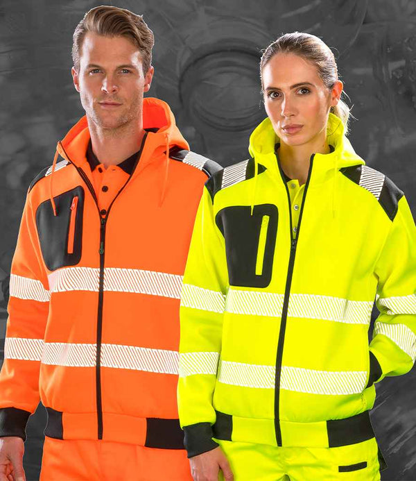 A man in an orange high-visibility Result Recycled Safety Zip Hoodie stands beside a woman in a yellow EN ISO 20471 jacket and pants, both featuring black accents and white reflective stripes for added safety.
