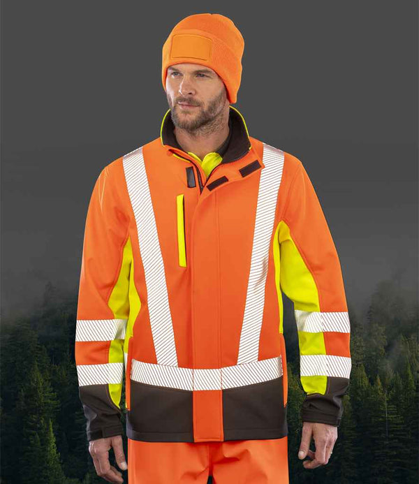 A man in a Result Recycled Three Layer Printable 3-Tone Safety Soft Shell Jacket and an orange beanie stands against a dark, blurred forest background.