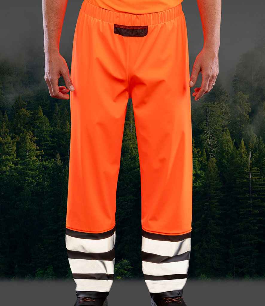 A person is shown from the waist down, wearing Result Recycled Heavy Duty Prism PU Safe and Dry Trousers by Result Recycled in bright orange with black and white reflective ankle stripes, standing in front of a dense forest with lush green trees.