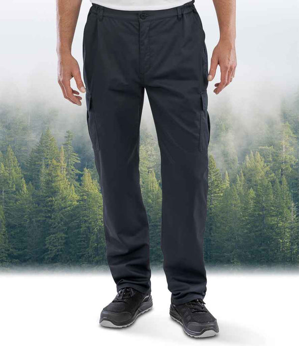 Someone wearing Result Recycled Work-Guard Utility Trousers from Result Recycled and black sneakers stands before a misty evergreen forest backdrop; only their lower body is visible.