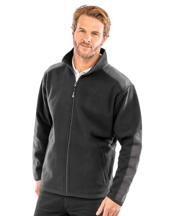 A man with light brown hair and a short beard wears the Result Recycled Heavy Duty Micro Fleece Jacket by Result Recycled. The black jacket features dark shoulder and arm panels. He stands against a white background, facing slightly left.