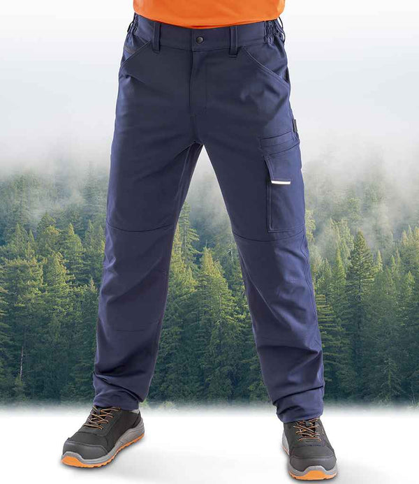 Wearing Result Recycled Dynamic Stretch Trousers in navy blue with articulated knees by Result Recycled, paired with gray and orange hiking shoes and an orange shirt, a person stands against a misty forest backdrop. The image shows only the lower body.