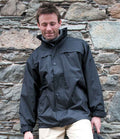A man in a Result Multi-Function Midweight Jacket by Result, featuring a black windproof design and adjustable hood, stands before a textured stone wall, looking down with a slight smile and eyes closed.