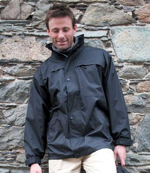 A man in a Result Multi-Function Midweight Jacket by Result, featuring a black windproof design and adjustable hood, stands before a textured stone wall, looking down with a slight smile and eyes closed.