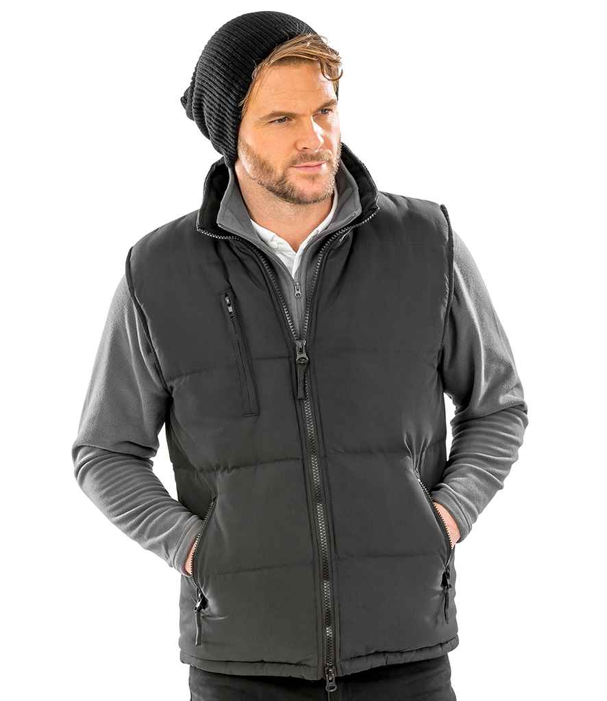 A man wearing a black beanie, grey long-sleeve shirt, and the Result Ultra Padded Bodywarmer by Result stands with his hands in his vest pockets, looking to the side against a plain white background.