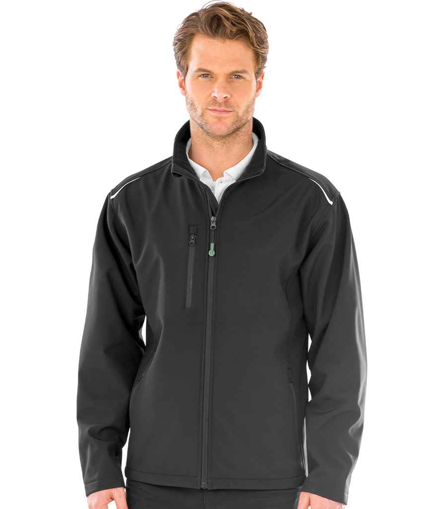 A man faces forward in a Result Recycled Three Layer Printable Soft Shell Jacket by Result Recycled—black, zip-up, waterproof with a high collar and chest pocket, made from recycled polyester—worn over a white collared shirt against a white background.