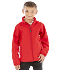 A young boy with short brown hair and a slight smile faces forward, wearing the Result Recycled Kids Two Layer Printable Soft Shell Jacket by Result Recycled in bright red, paired with black pants. The background is plain white.