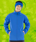 A person wears a Result Recycled Printable Soft Shell Jacket by Result Recycled and a blue-and-black beanie, standing with hands in pockets against a bright green abstract background.